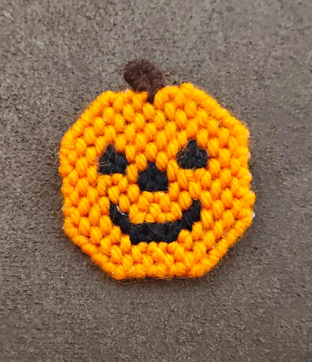 Orange crocheted jack o' lantern face brooch with black details and brown fuzzy stem.