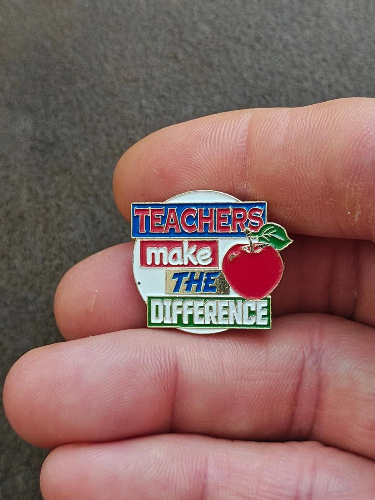 Enamel teacher pin held between fingers, red apple with green leaf and slogan