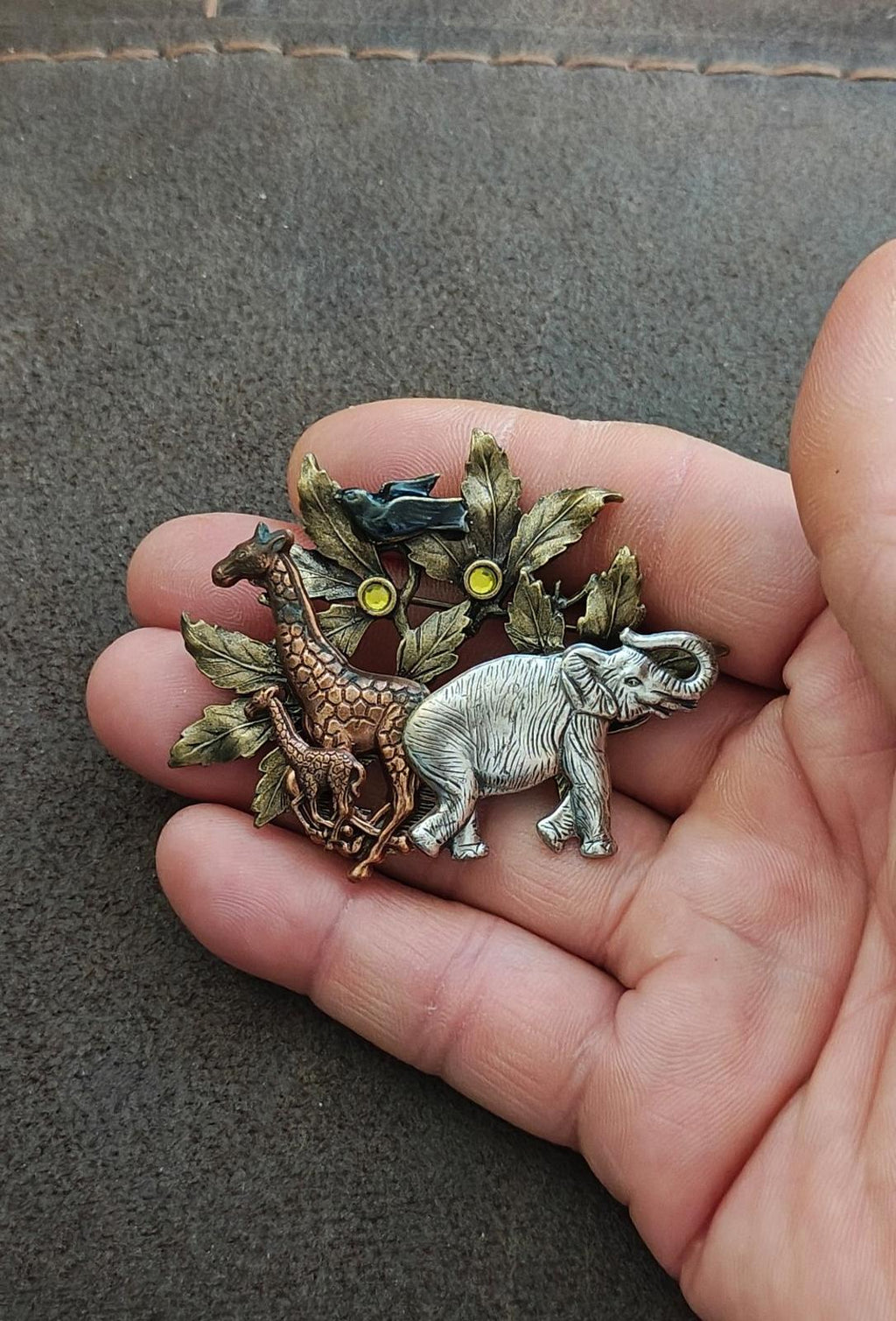 Safari scene brooch in hand: silver-tone elephant, leafy branches, and copper-toned giraffe figure