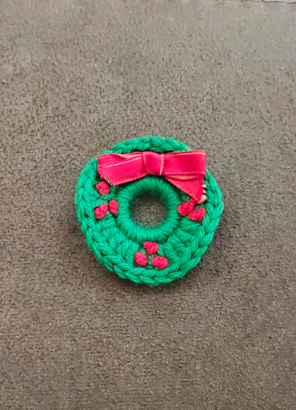 Green knitted wreath brooch with pink bow and red berry accents, flat round design
