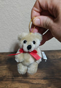 Handheld beige plush teddy bear ornament with red bow and gold loop cord