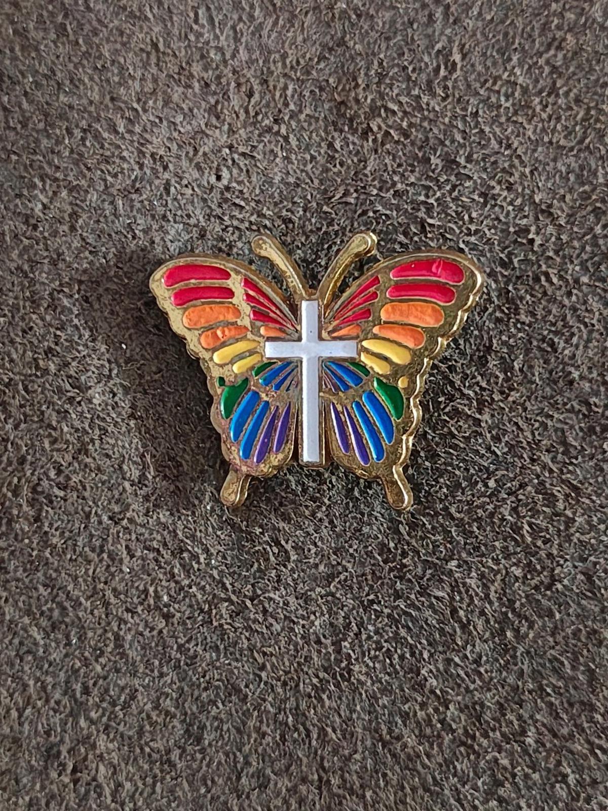 Rainbow butterfly enamel pin with red, blue, and yellow wings plus white cross