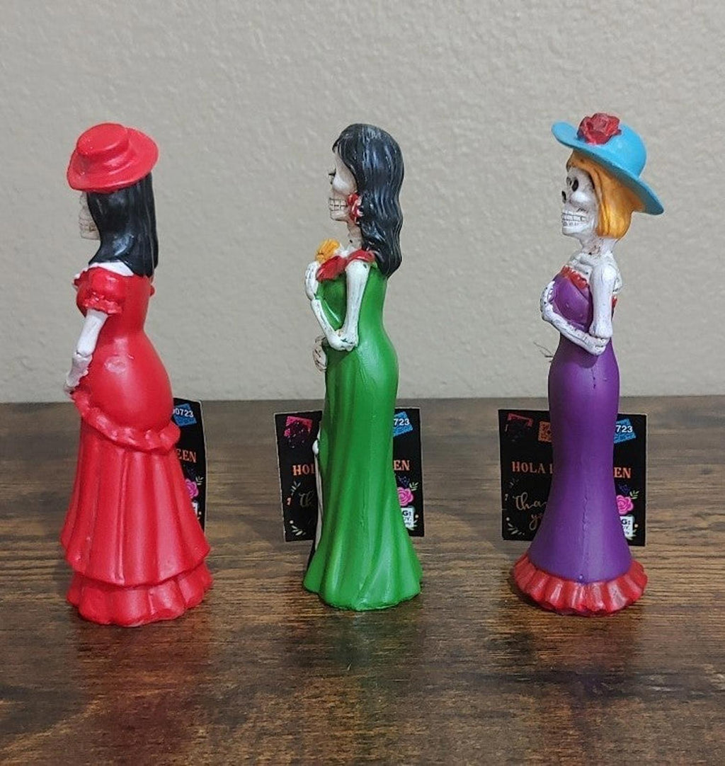 Three Catrina figurines in side view, red, green, and purple dresses
