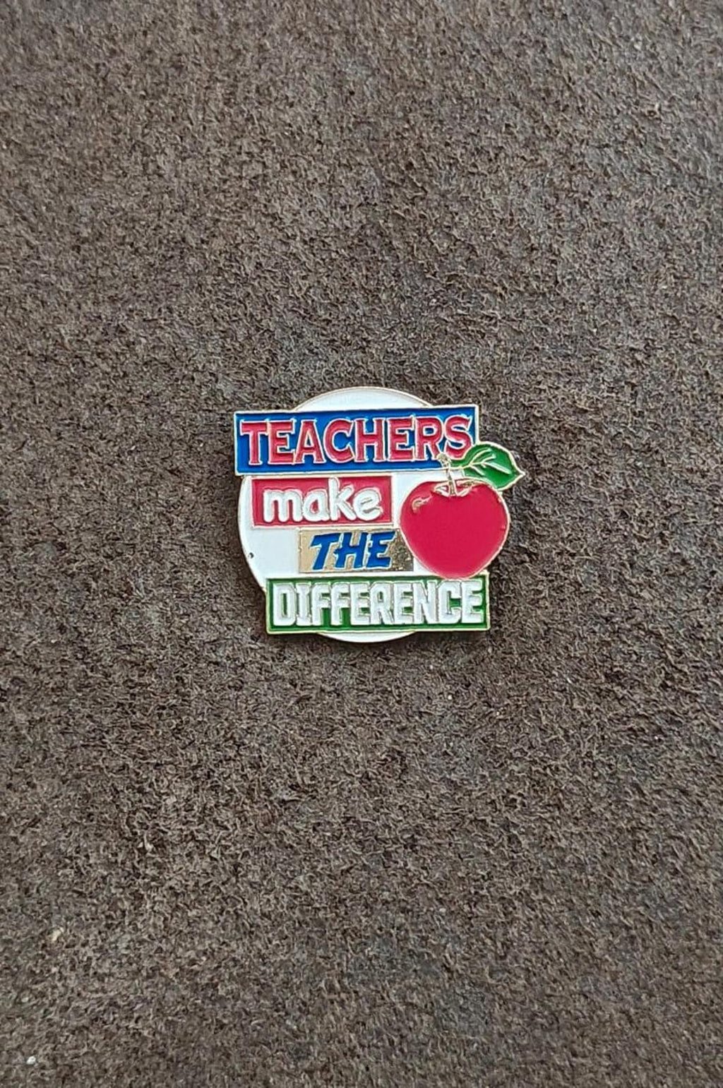 Enamel teacher pin on gray surface, red apple and “Teachers make the difference” text