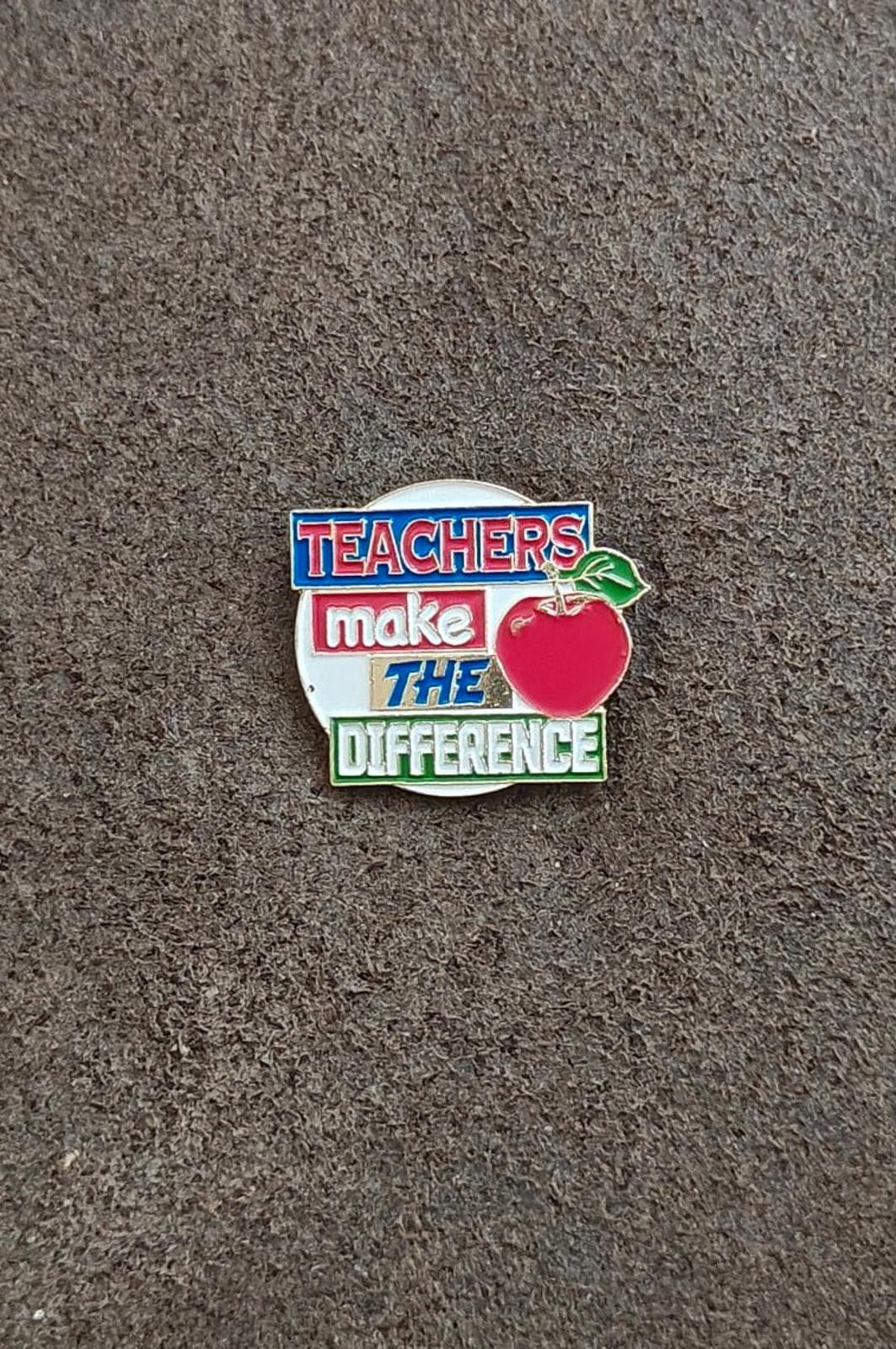 Enamel teacher pin on gray surface, red apple and “Teachers make the difference” text