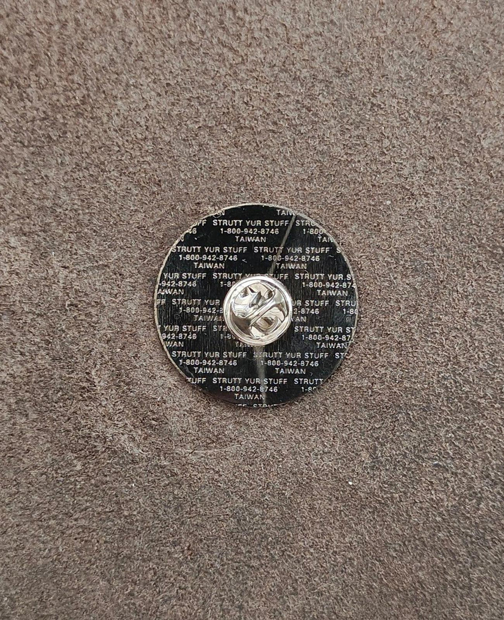 Back of gold tone lapel pin with stamped text and circular clasp detail.