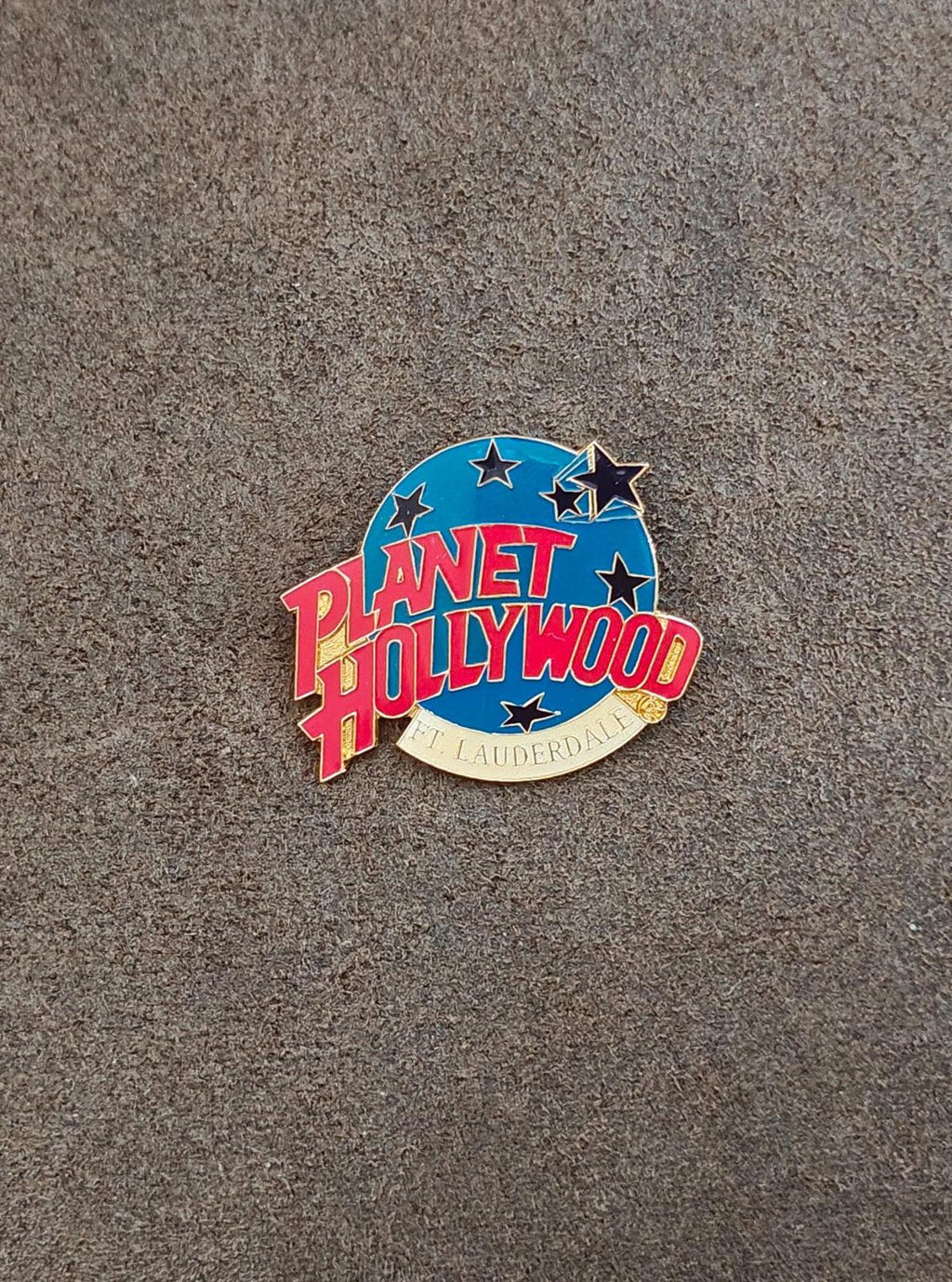 Blue enamel lapel pin with red Planet Hollywood lettering and stars, gold backing.