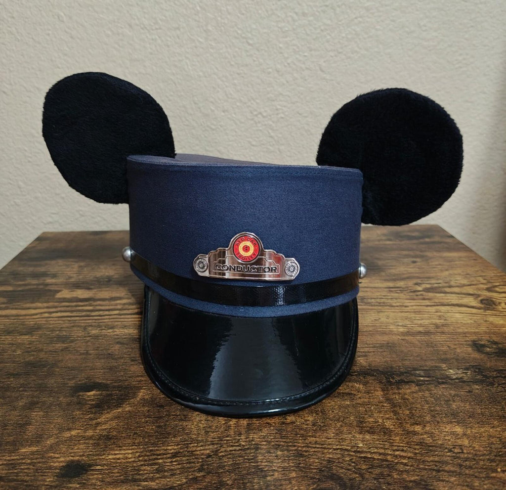 Front view of blue conductor hat with black Mickey ears and red-and-gold conductor badge.