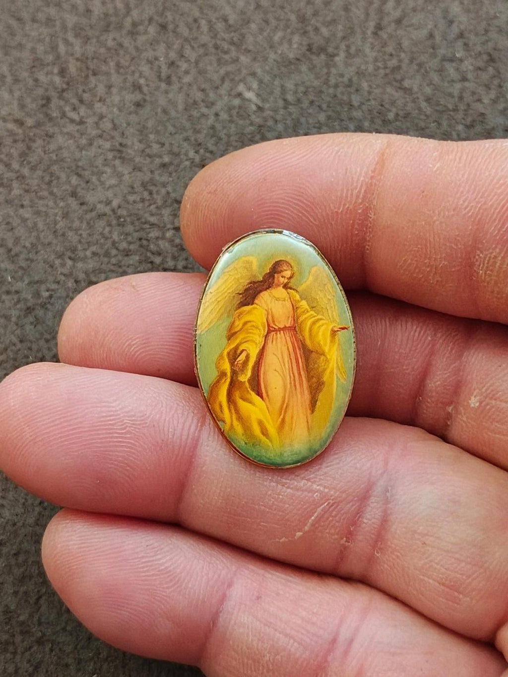 Small oval guardian angel pin in lucite-style frame, shown on fingertip close-up