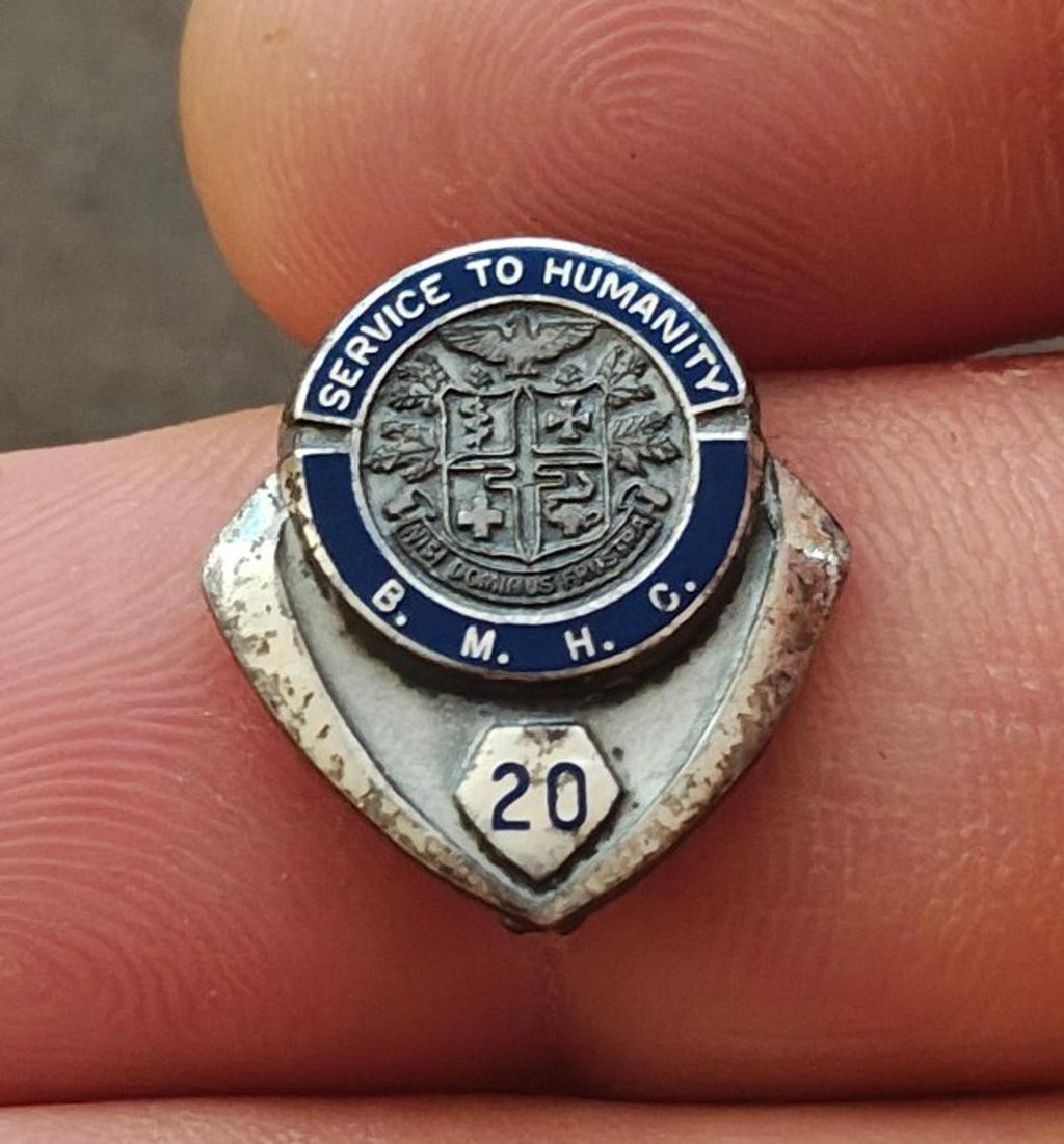 Silver enamel service award pin close-up with “Service to Humanity” shield and “20”.
