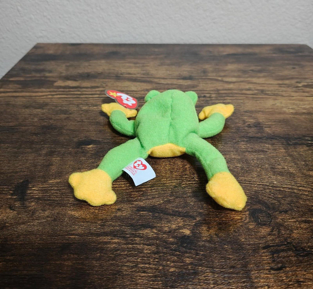 Green frog beanbag plush lying on wooden tabletop with red swing tags