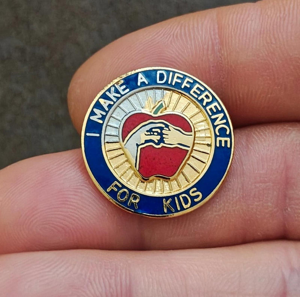 Close-up gold-tone enamel pin with blue ring text “Make a Difference for Kids” and apple icon.