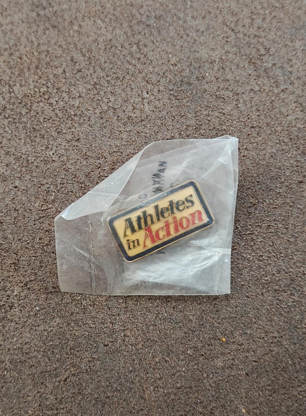 Gold-tone enamel "Athletes in Action" pin in clear plastic bag on gray surface