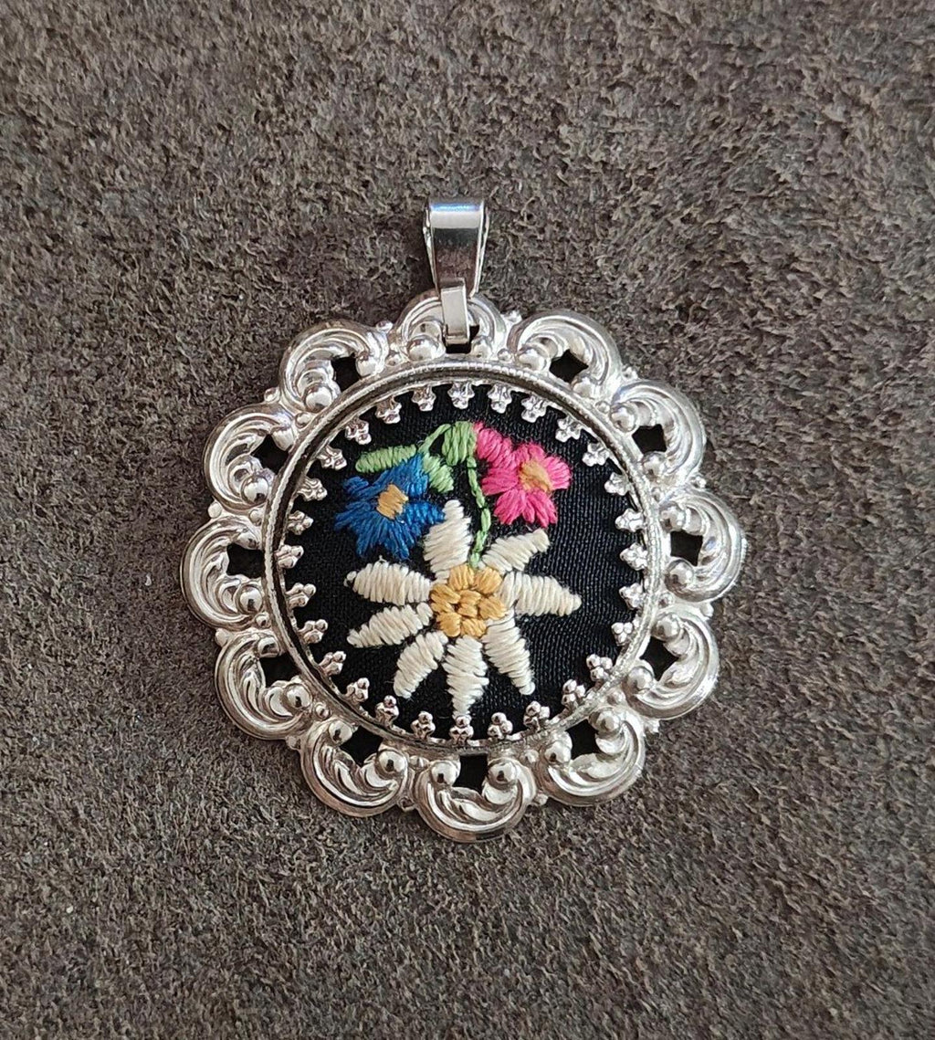 Silver filigree pendant with round black embroidered flower and decorative side scrolls.