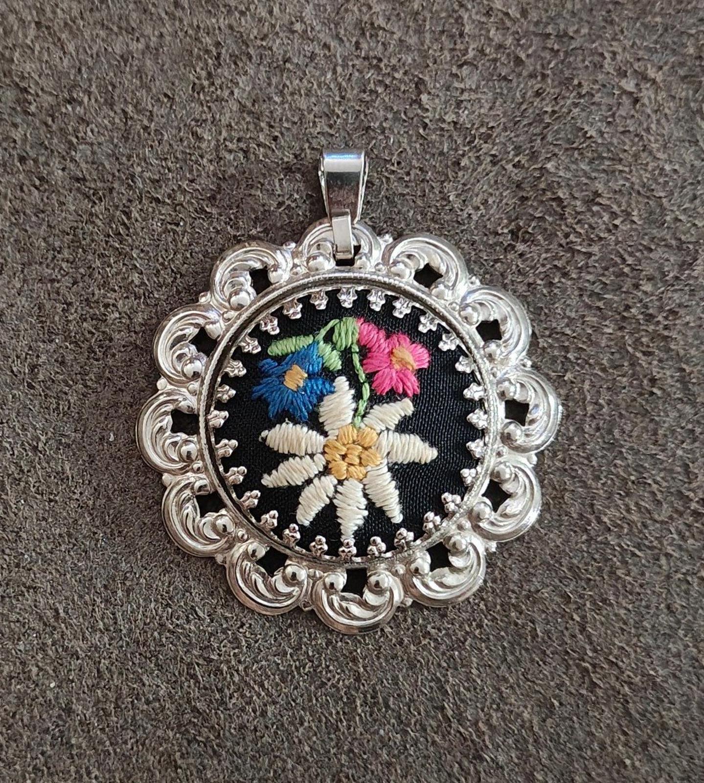 Silver filigree pendant with round black embroidered flower and decorative side scrolls.