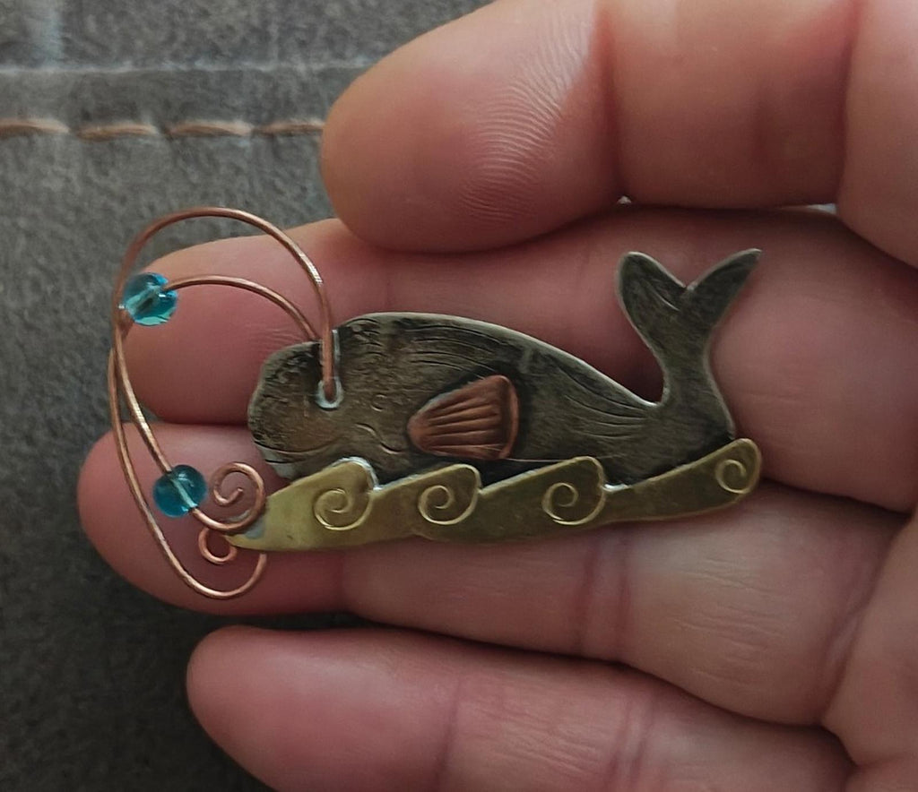 Gold-tone whale brooch pin closeup with blue stones, copper wire swirl, dark green enamel whale