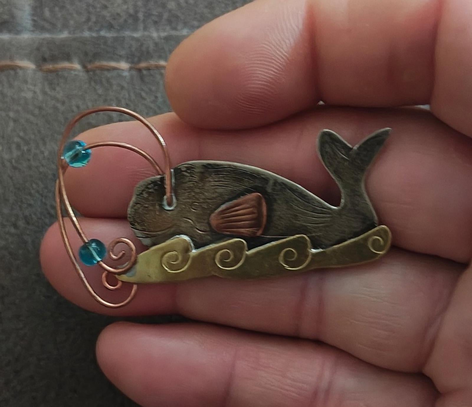 Gold-tone whale brooch pin closeup with blue stones, copper wire swirl, dark green enamel whale