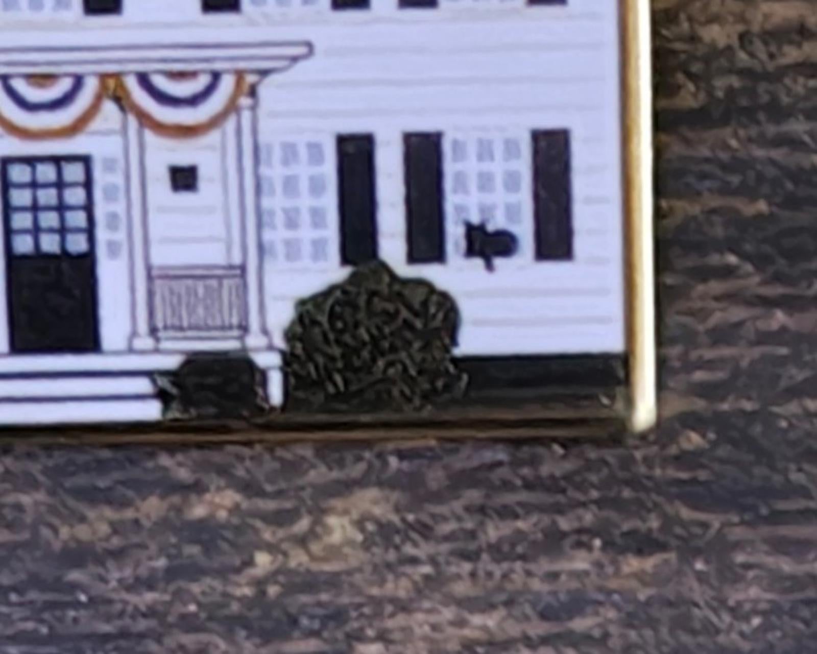 Close-up of enamel pin showing entrance area and black shrub silhouettes, gold border