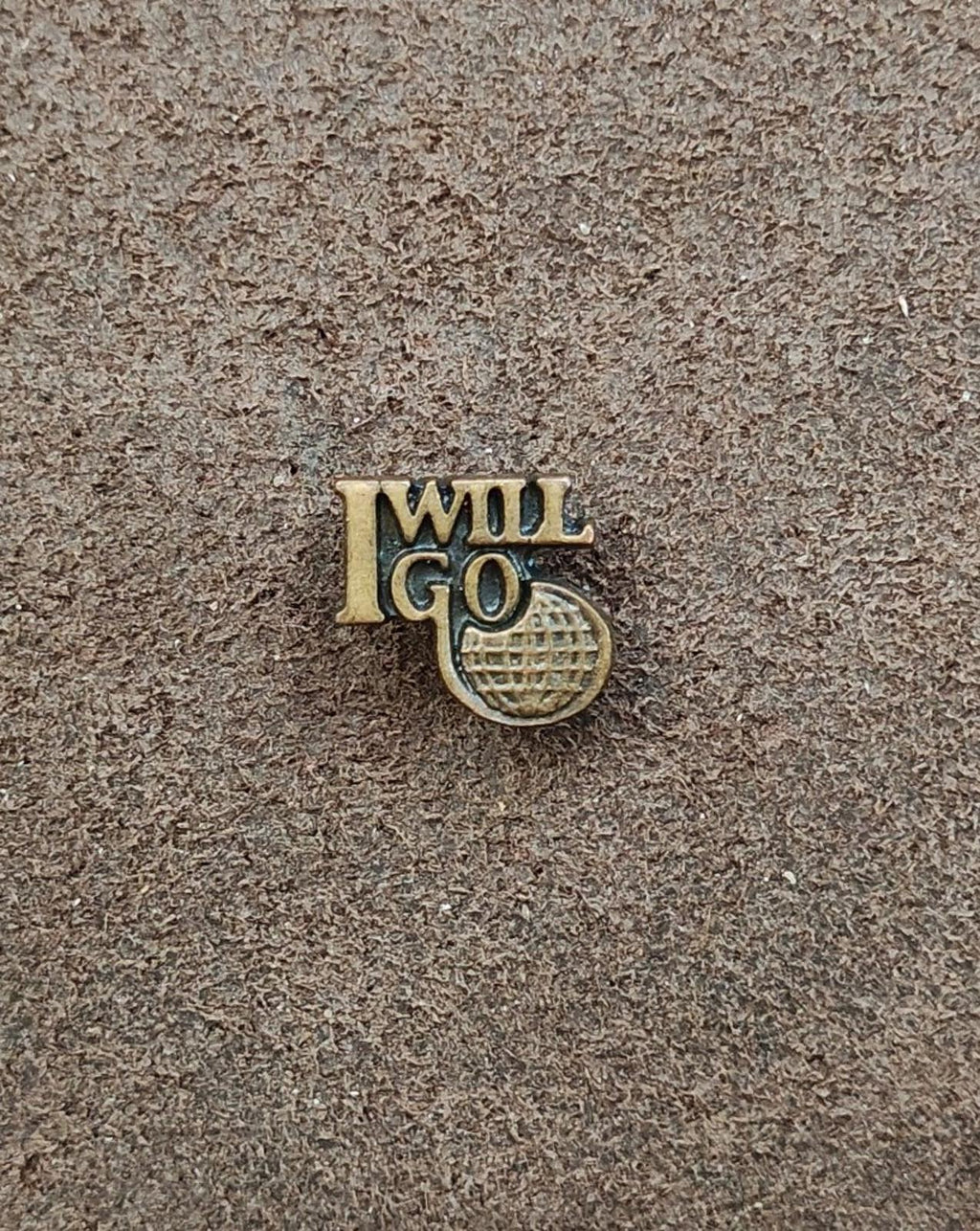 Gold tone "I WILL GO" lapel pin with globe-style emblem, flat on textured surface