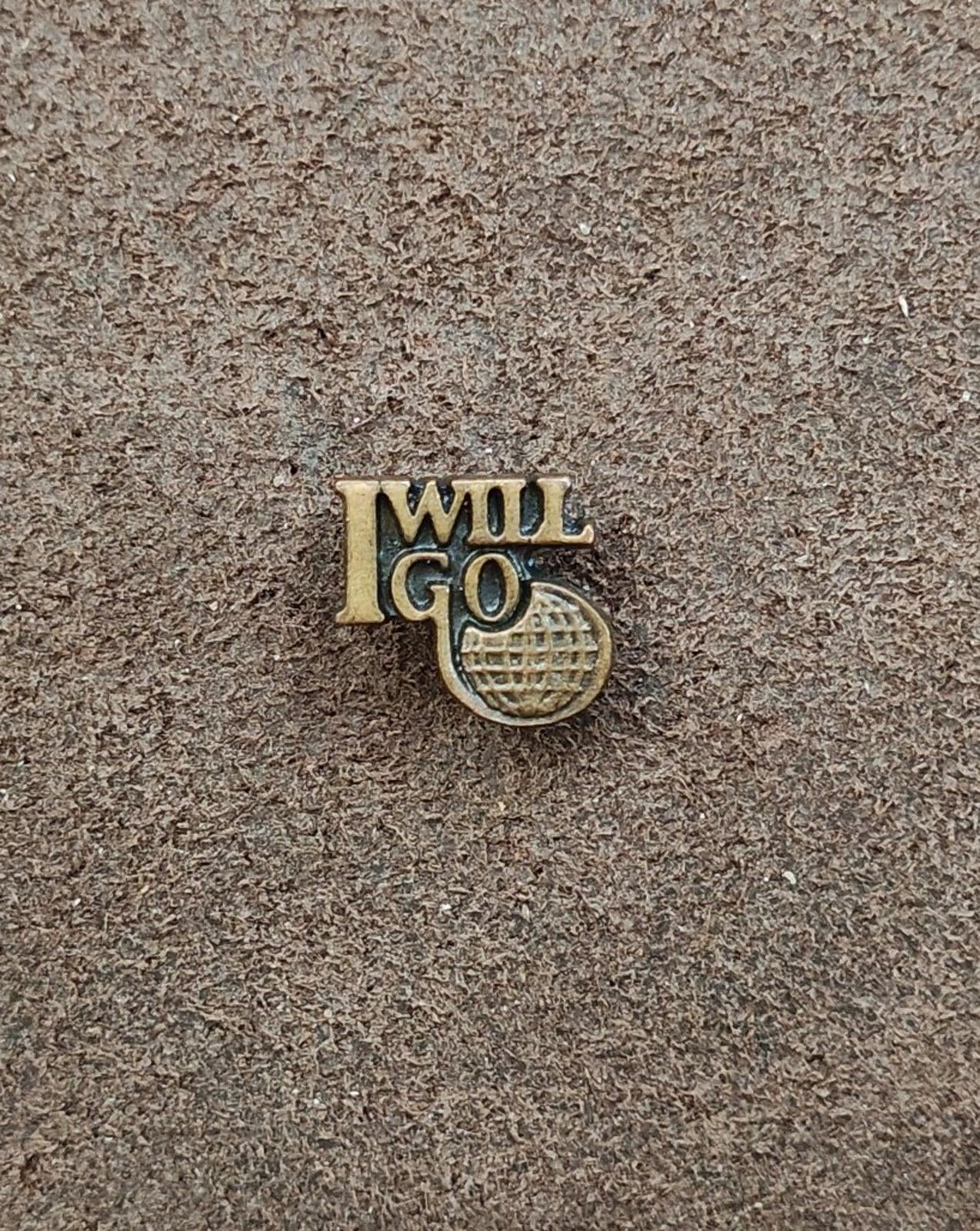 Gold tone "I WILL GO" lapel pin with globe-style emblem, flat on textured surface