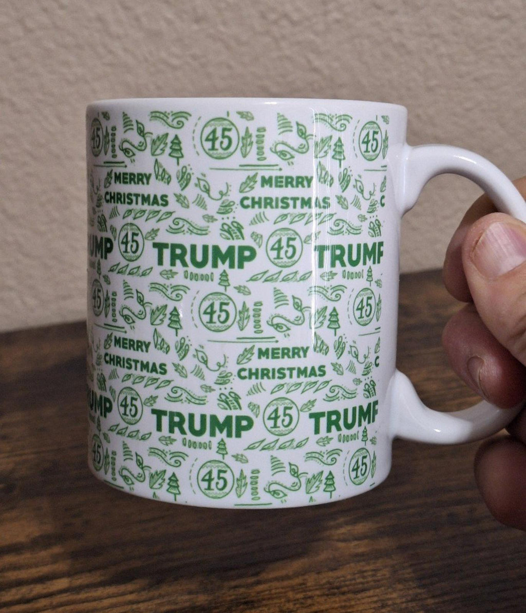 White ceramic mug with green “MERRY CHRISTMAS” and “TRUMP” text and circular 45 icons.