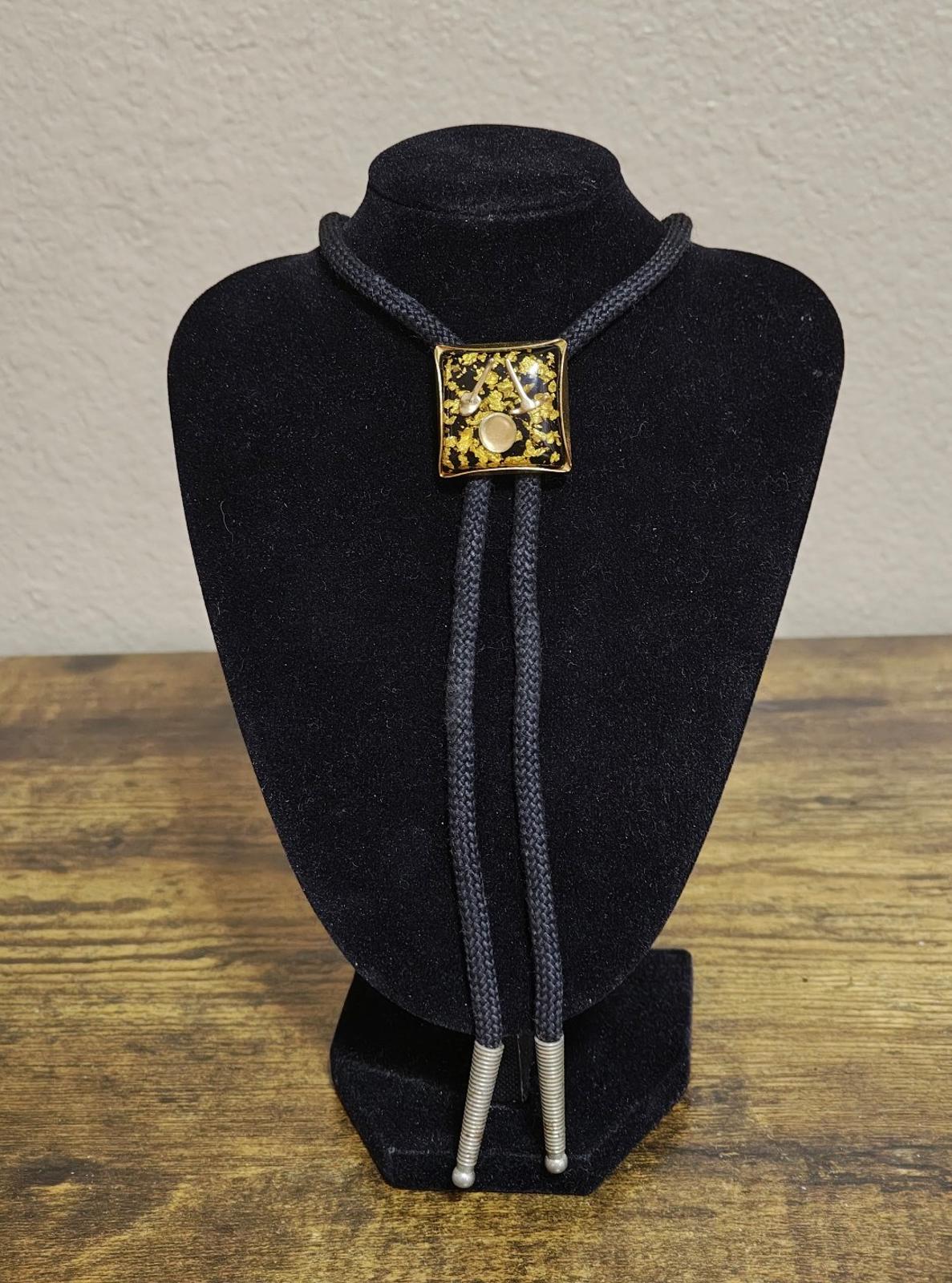 Black bolo tie with gold square pendant and twin cords on stand