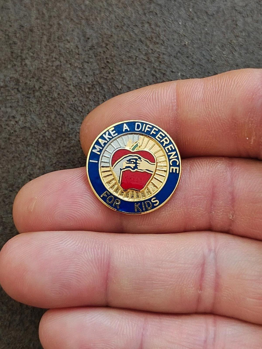 Small round gold-tone enamel pin with blue text ring and red apple icon.