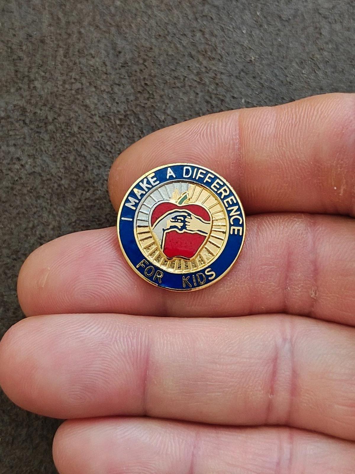 Small round gold-tone enamel pin with blue text ring and red apple icon.