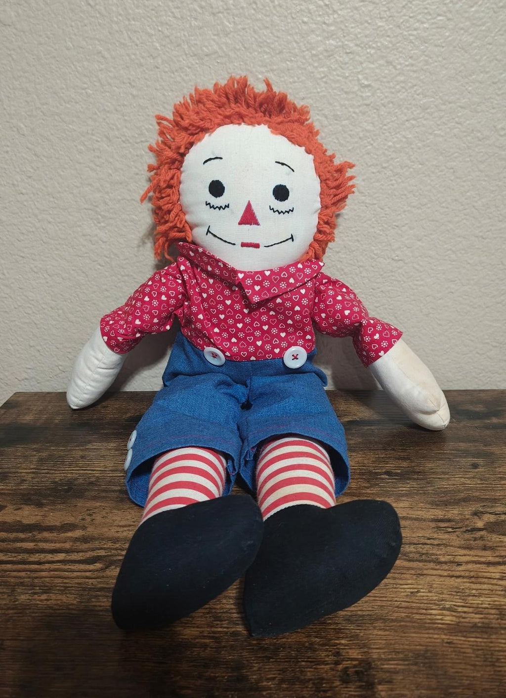 Handmade rag doll seated, orange yarn hair, red heart-pattern shirt, blue overalls, striped legs.