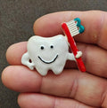 Tooth-shaped enamel pin holding a red toothbrush with teal handle, white tooth detail.