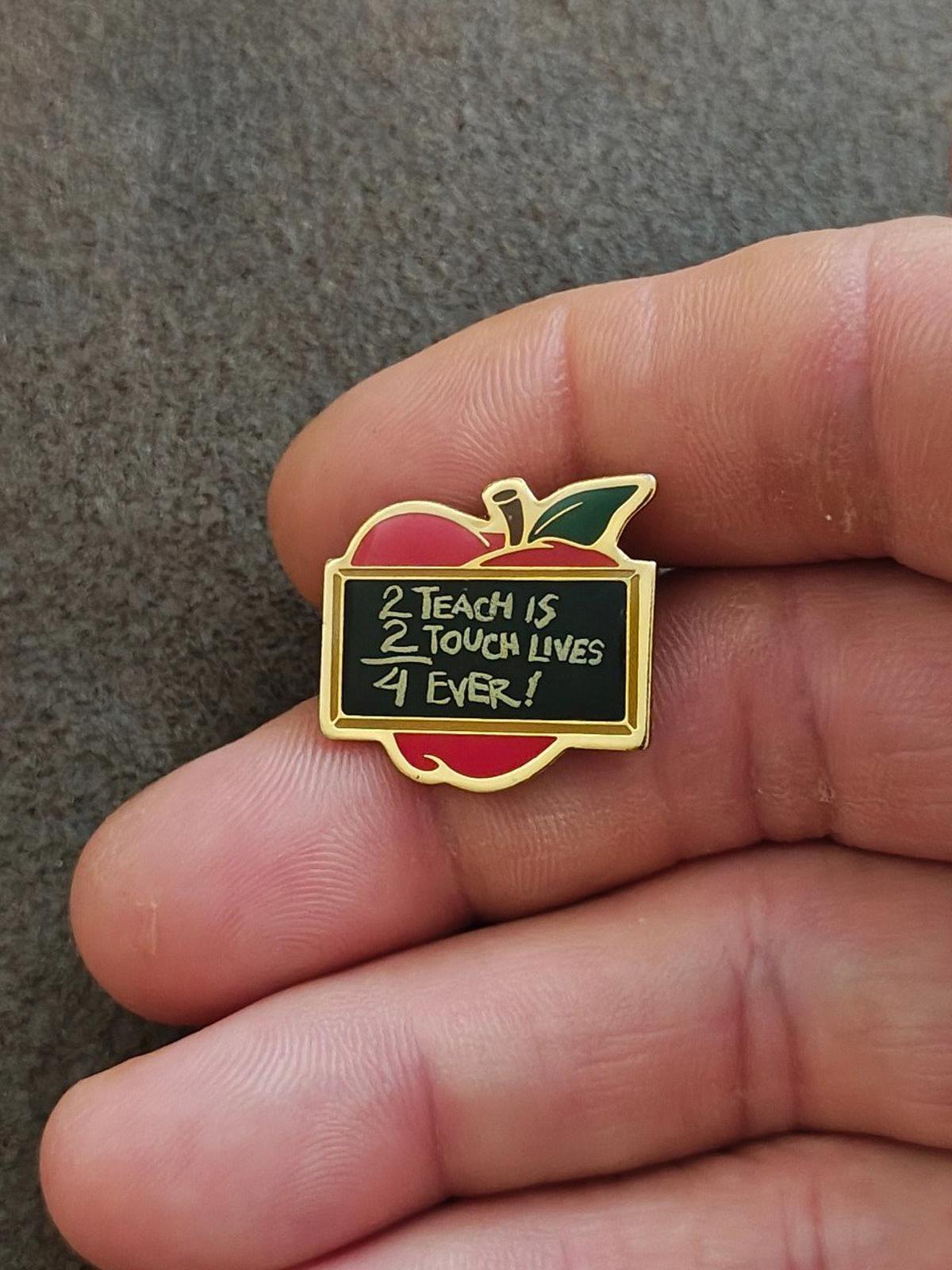 Gold-tone apple teacher pin with red enamel apple and green leaf, held close to camera