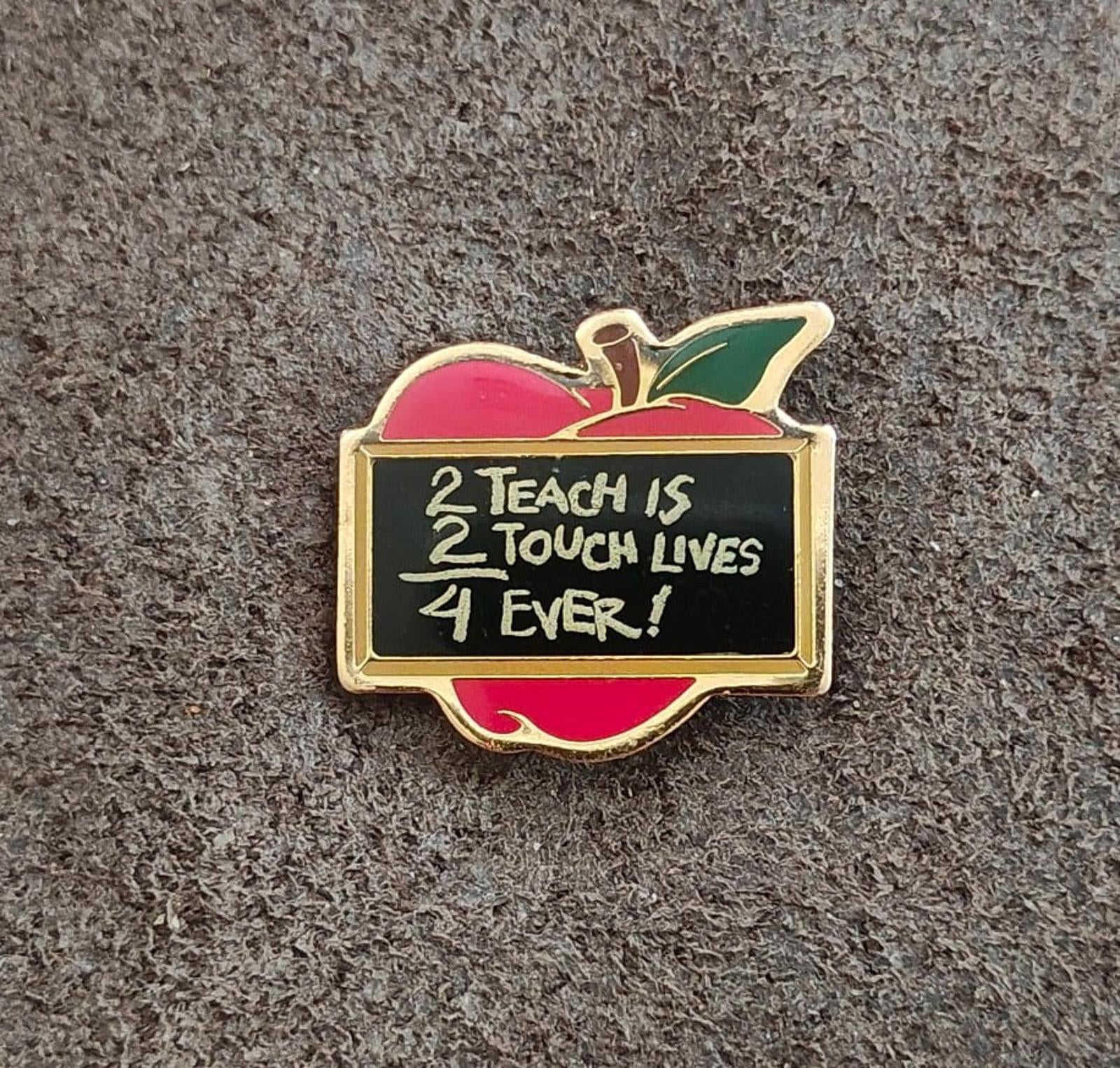 Gold-tone apple teacher pin with red base, green leaf, and “Teach is / Touch Lives / Ever!” text
