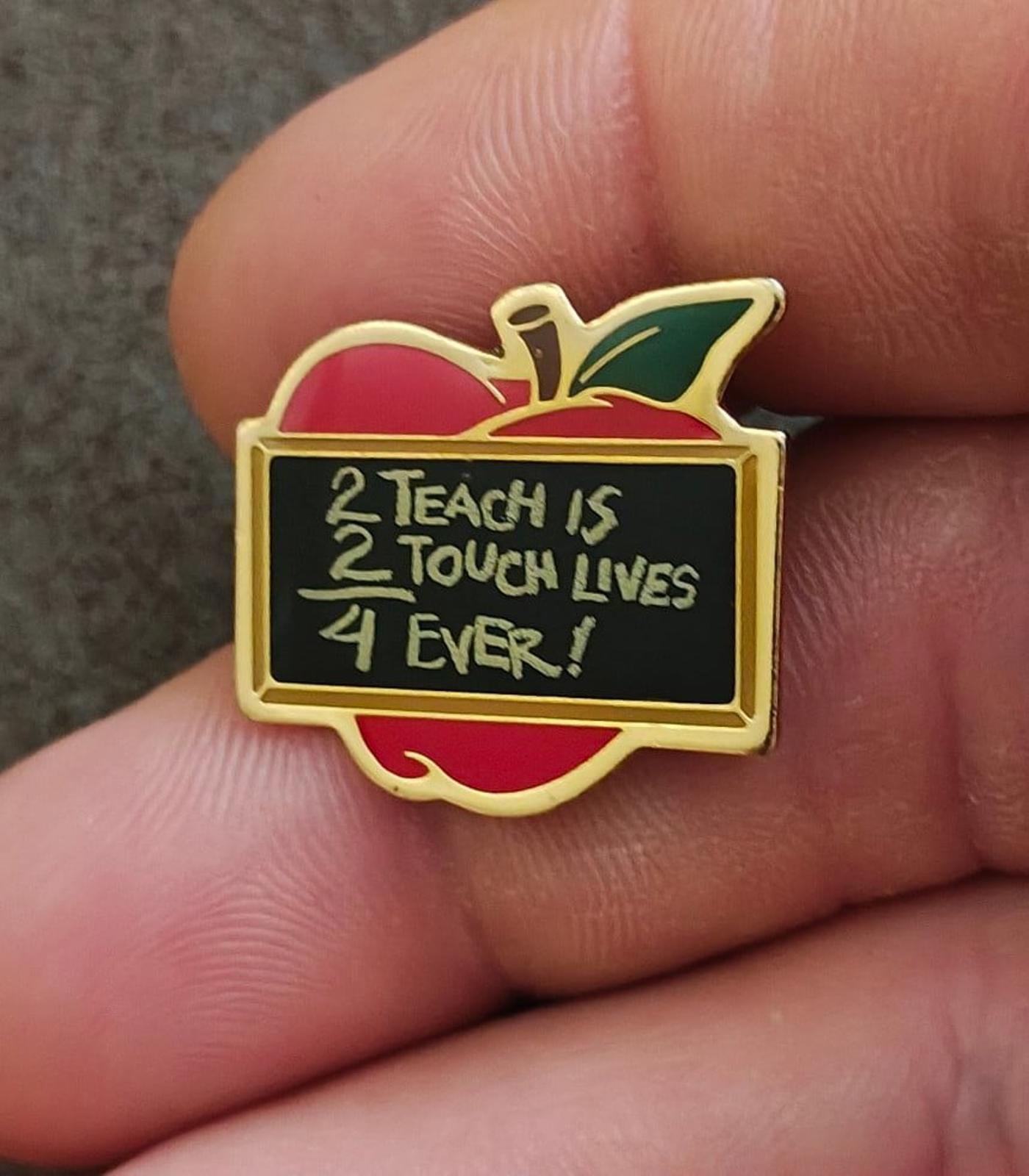 Gold-tone apple teacher pin with red and green enamel and black quote panel reading “2 Teach is…”