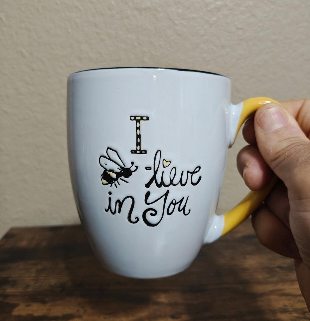 White inspirational mug with bee design and “live in You” text, held by yellow handle