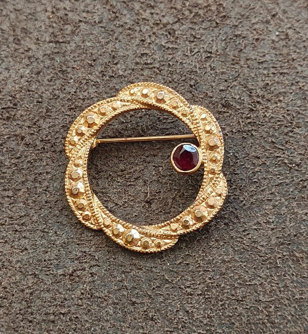 Gold tone circular brooch with dark red garnet set in an ornate beaded ring design.