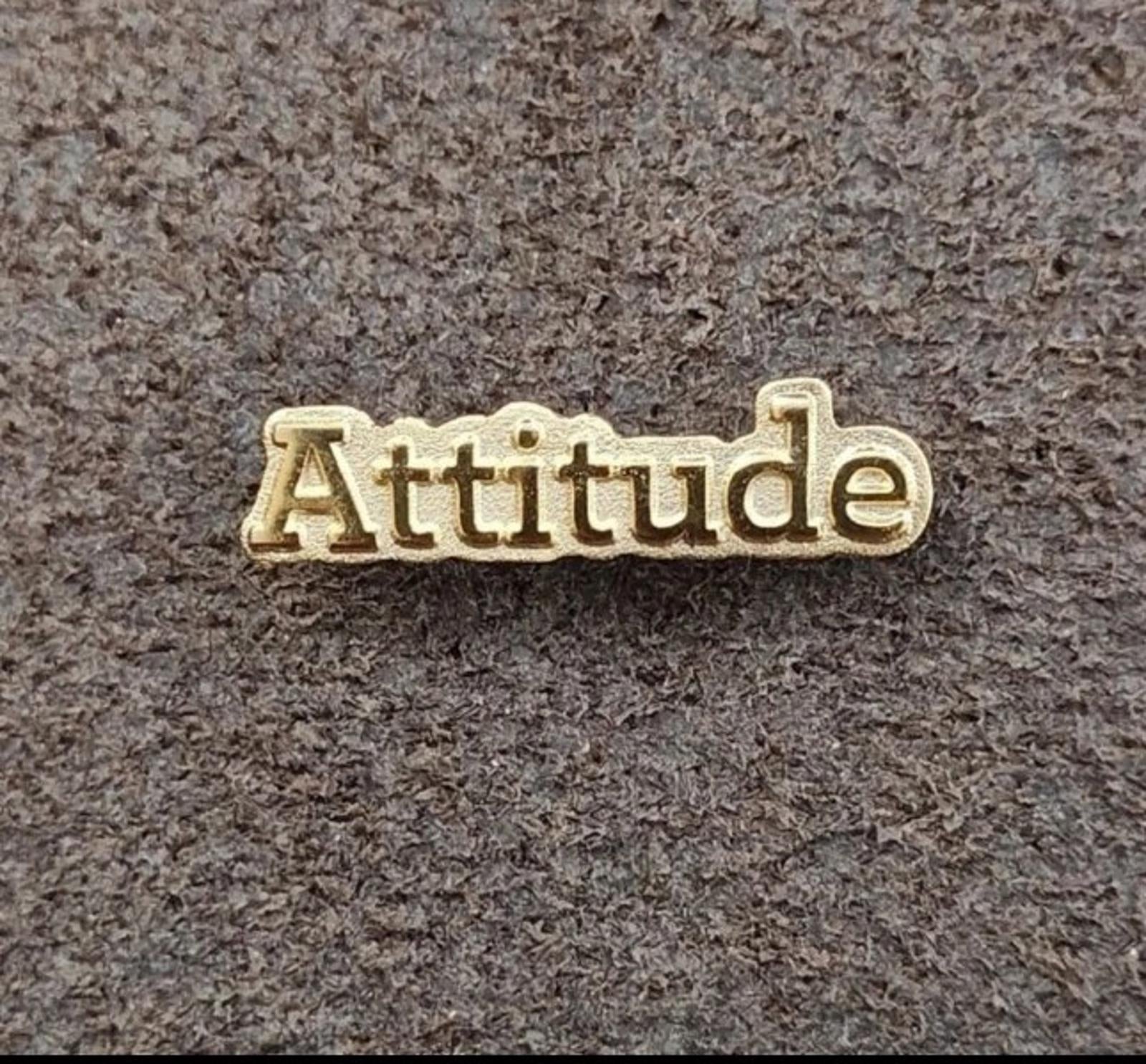 Gold tone lapel pin with raised "Attitude" lettering on textured gray surface