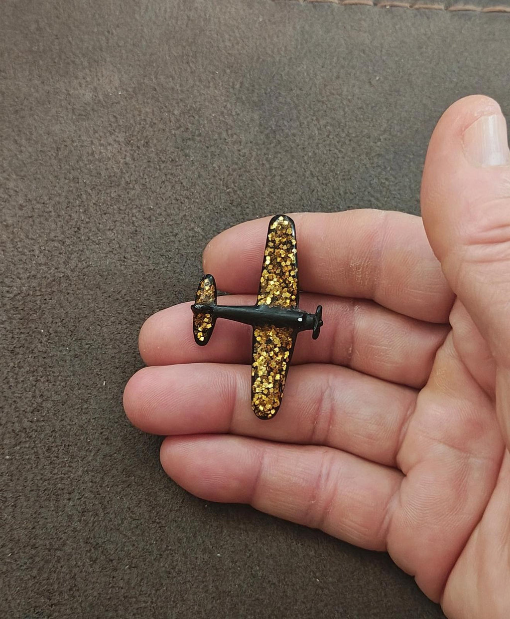 Airplane brooch in gold glitter with dark accents, held in open palm