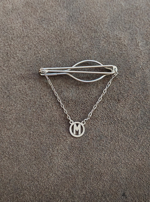 Silver tone M monogram pendant on chain beneath a bar clip with curved wire frame.