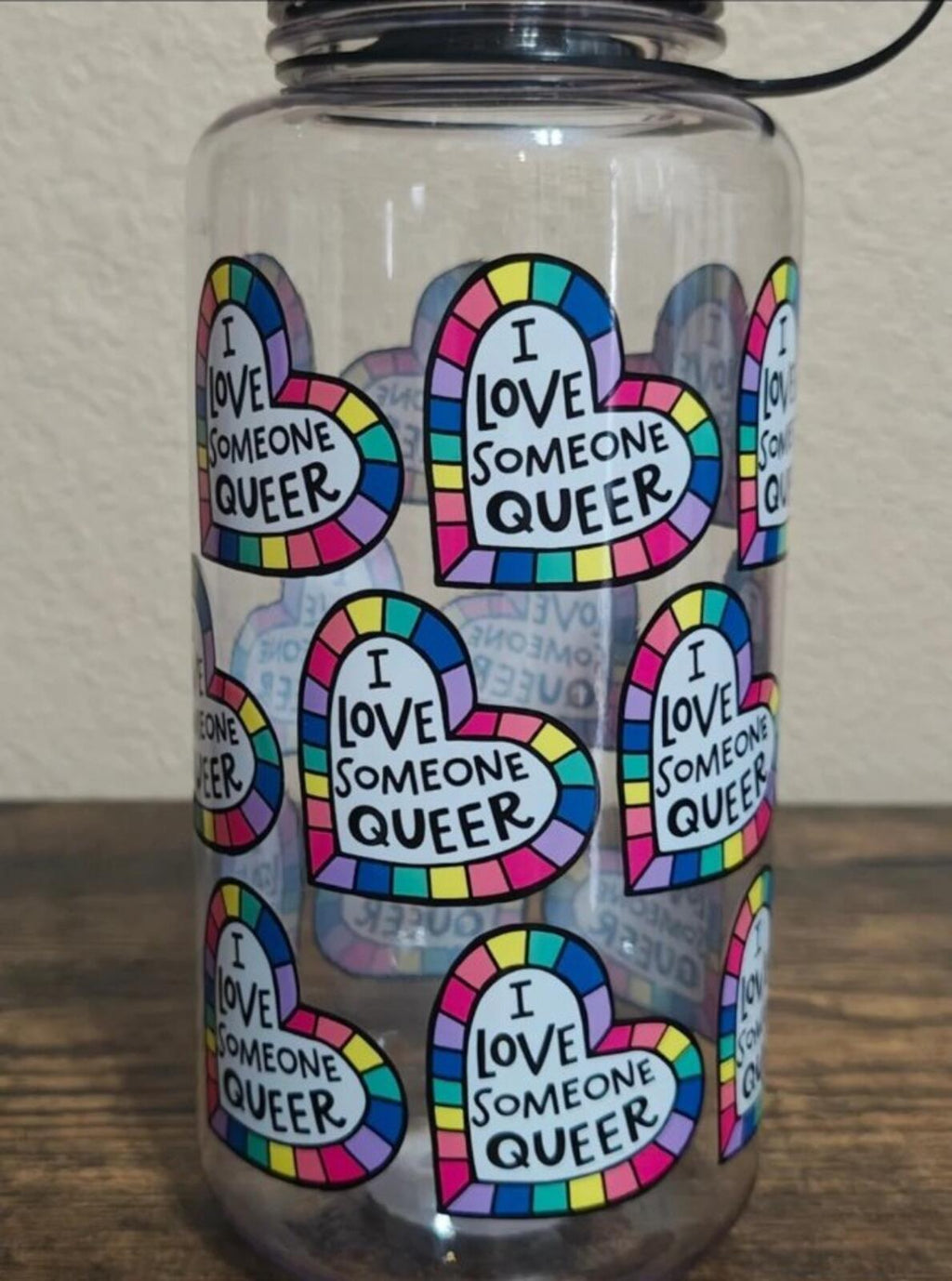 Clear water bottle with black lid, close-up of colorful 'I love someone queer' heart logos
