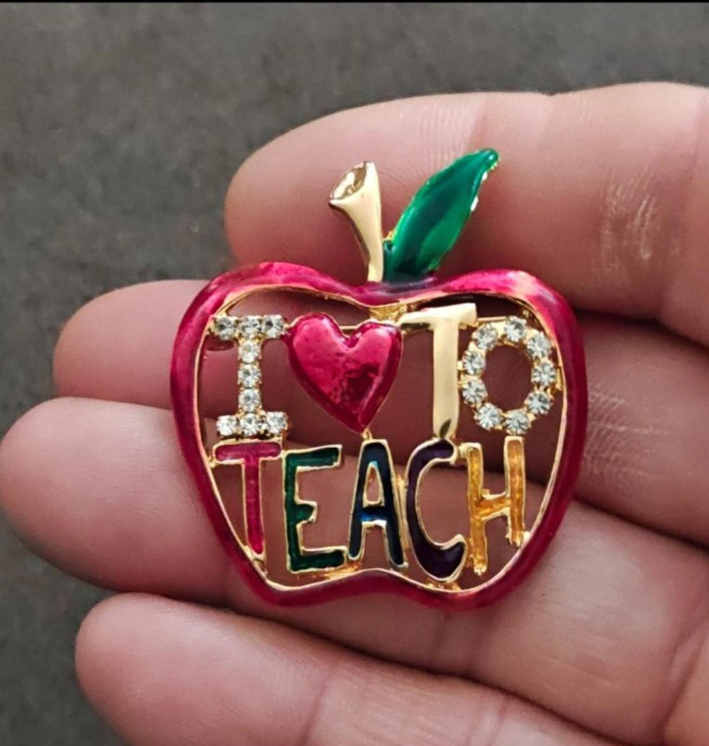 Red enamel apple-shaped brooch with gold tone frame, rhinestones, green leaf, and "TEACH" heart.