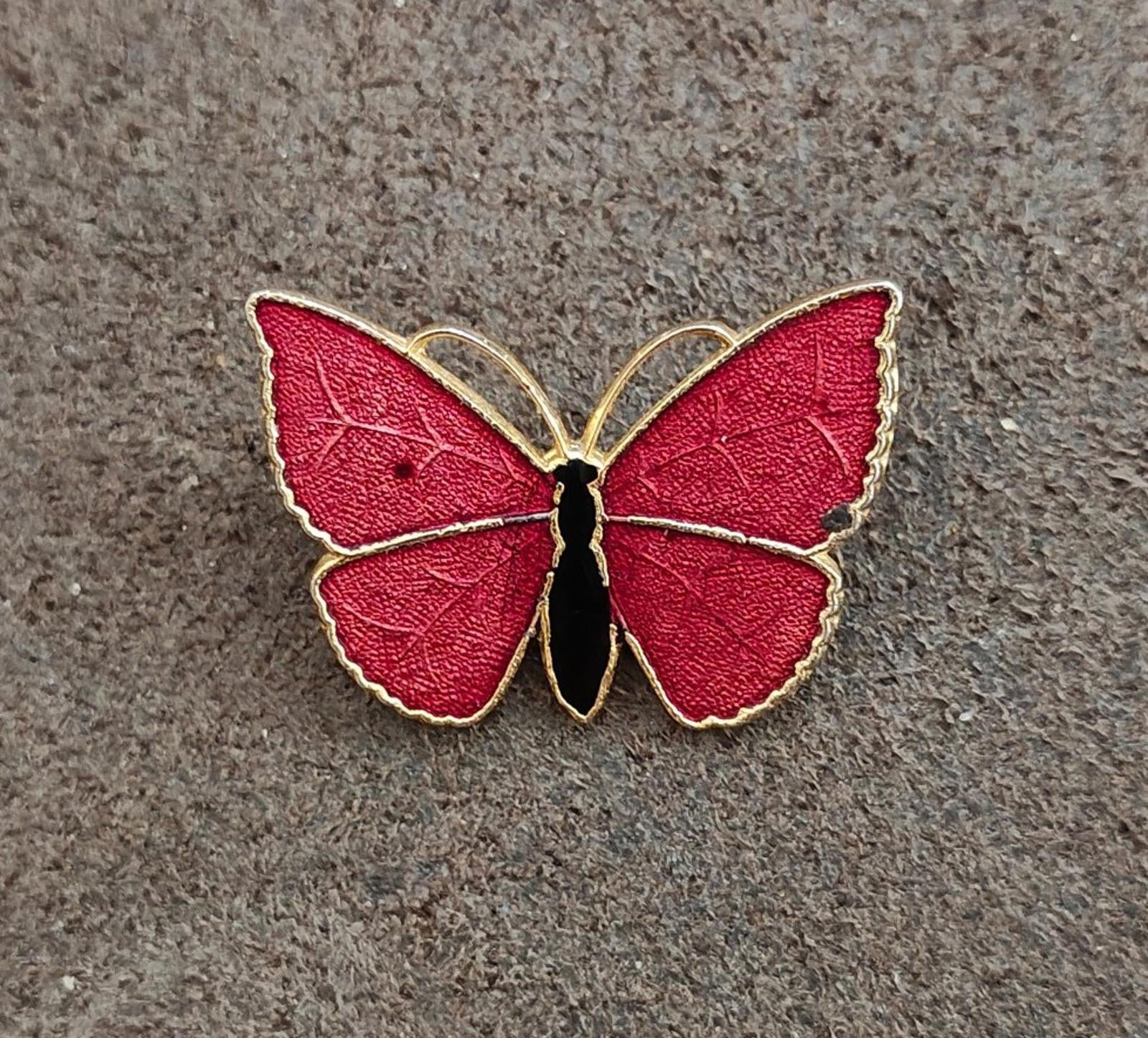 Red butterfly enamel brooch with gold-tone edging and black body, flat on textured surface.