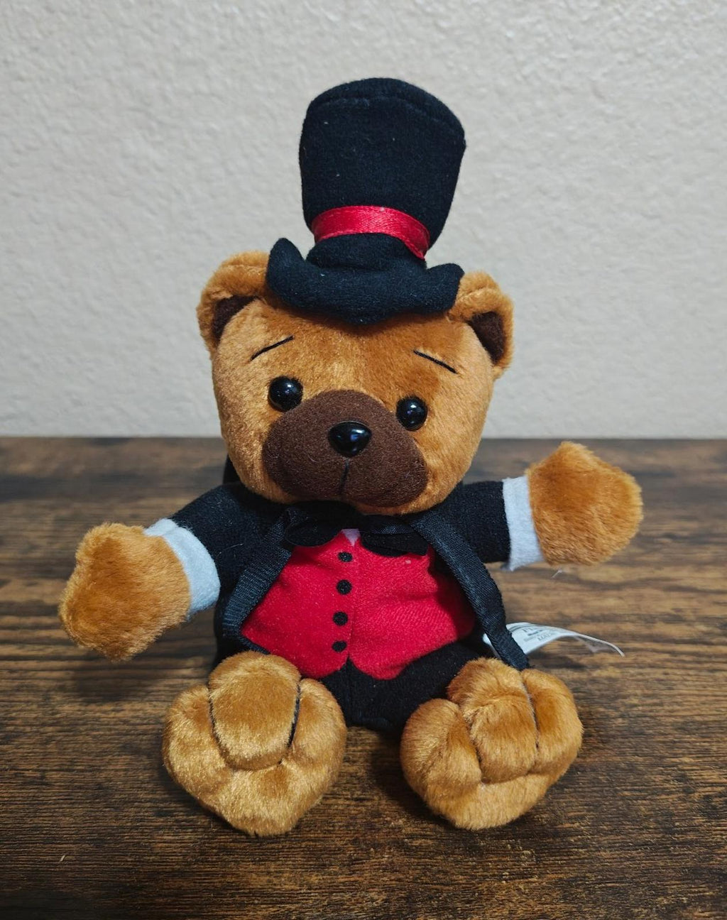 Plush bear seated on table wearing black tuxedo, red vest, black bow tie, top hat.