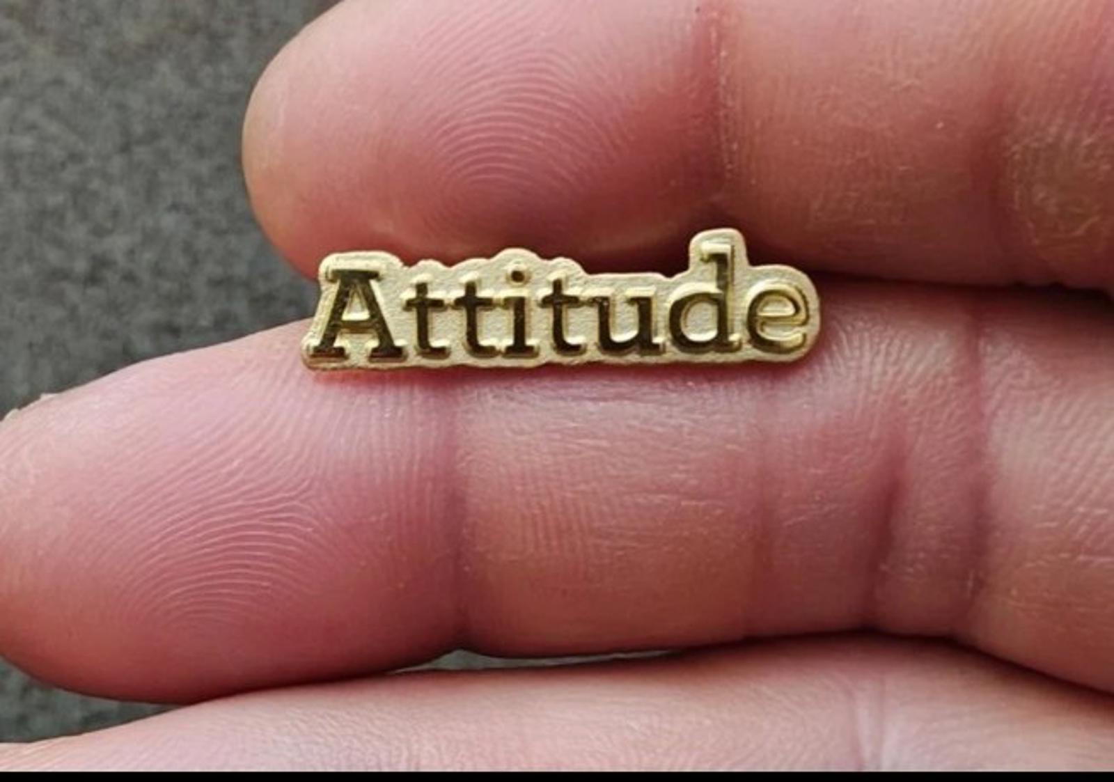 Gold tone lapel pin with raised "Attitude" lettering held between fingers, close-up