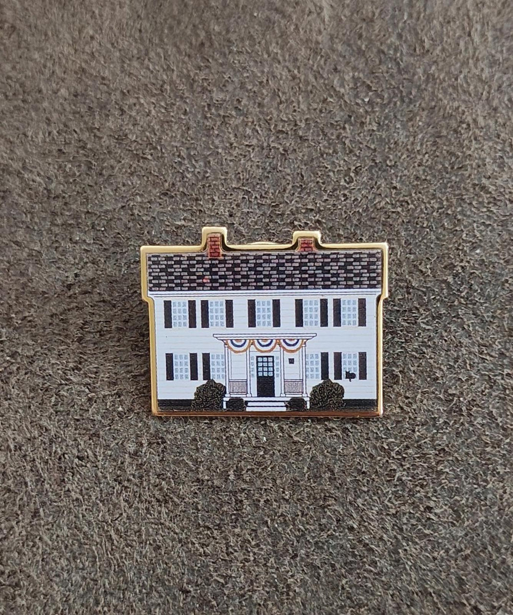 Enamel pin on gray fabric, front view of white building with blue windows and gold border