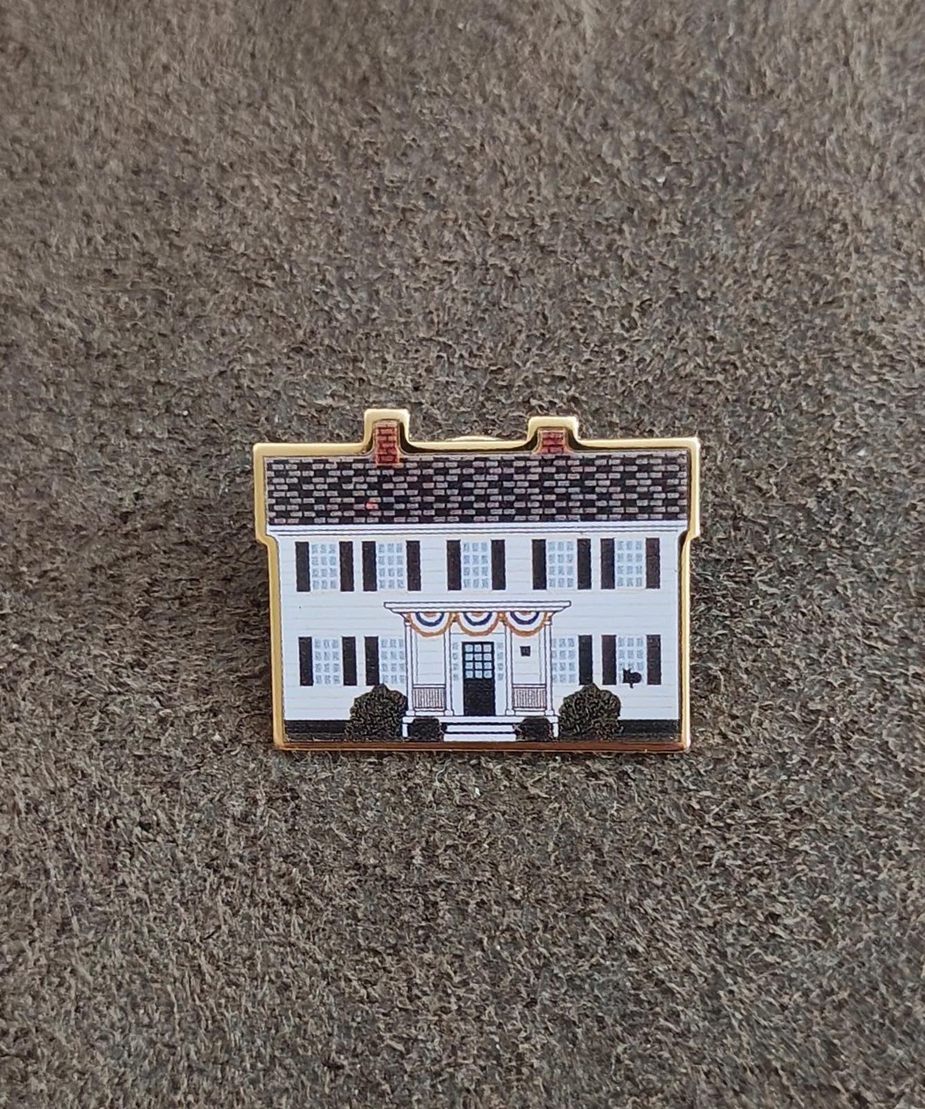Enamel pin on gray fabric, front view of white building with blue windows and gold border