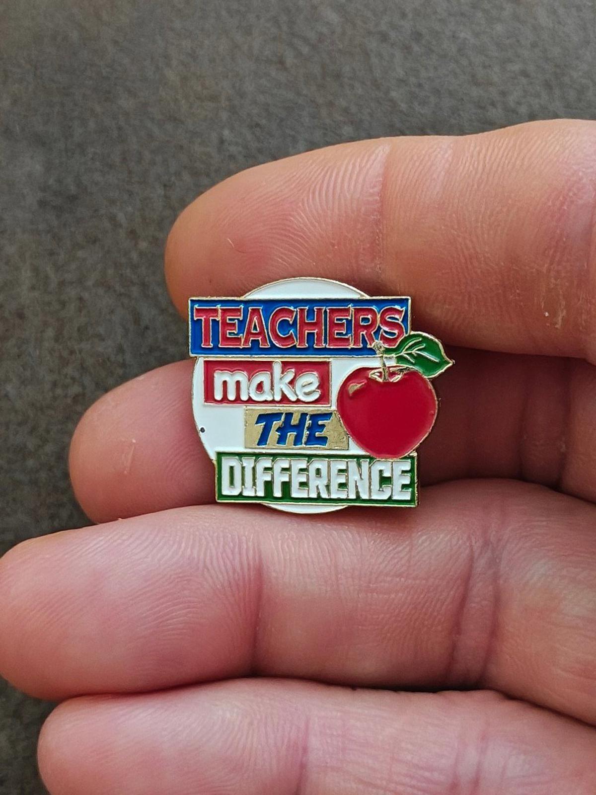 Enamel teacher pin close-up in hand, blue “TEACHERS” and red apple detail
