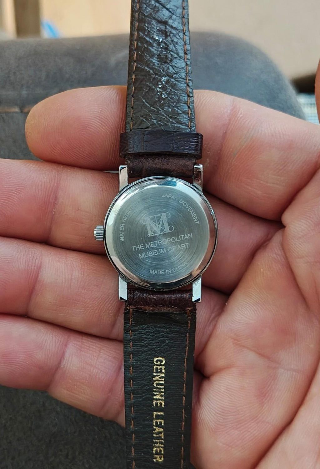 Round case wristwatch back with text, brown leather strap showing “GENUINE LEATHER”