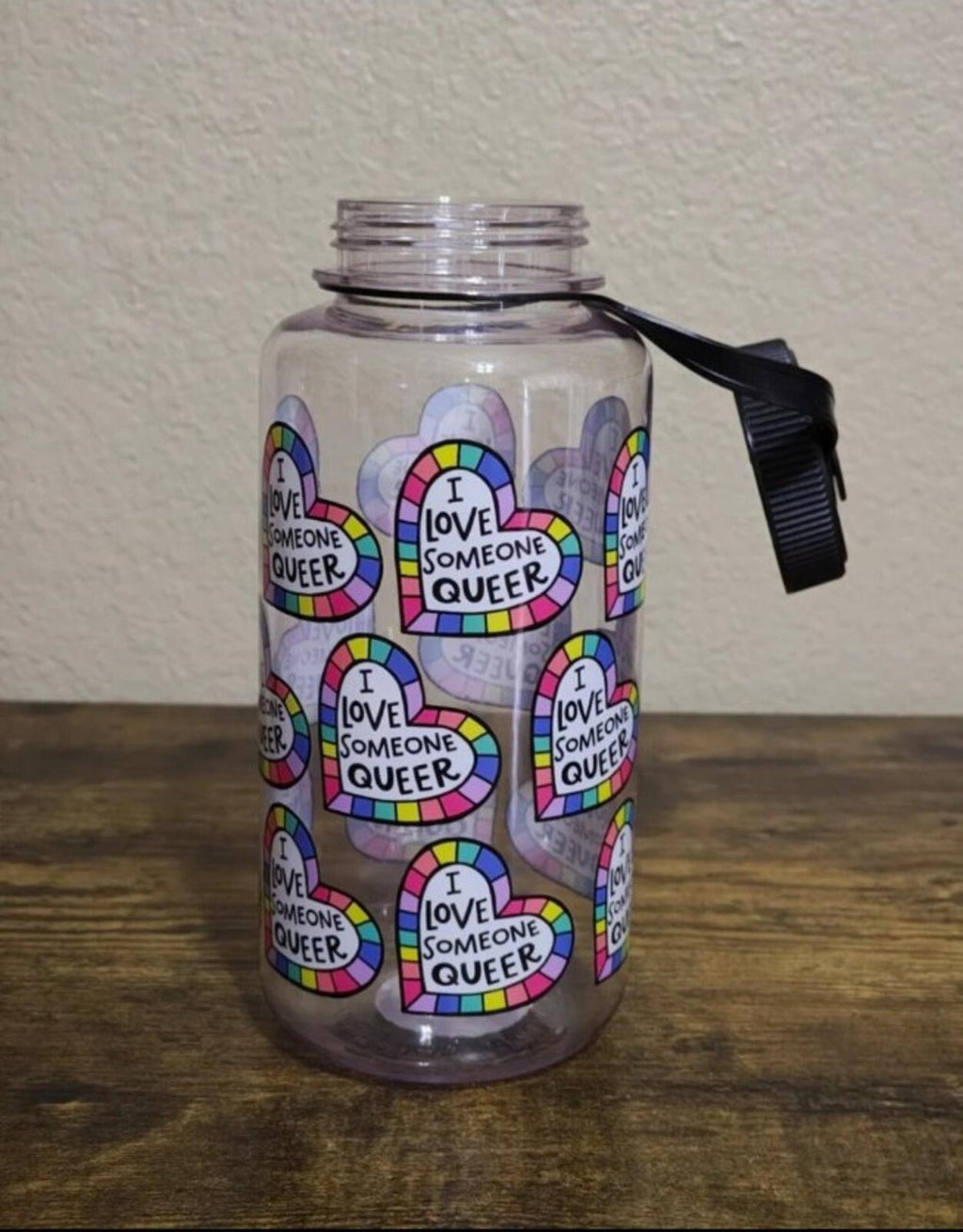 Clear 32 oz plastic water bottle with black lid and carry strap, colorful 'I love someone queer' hearts