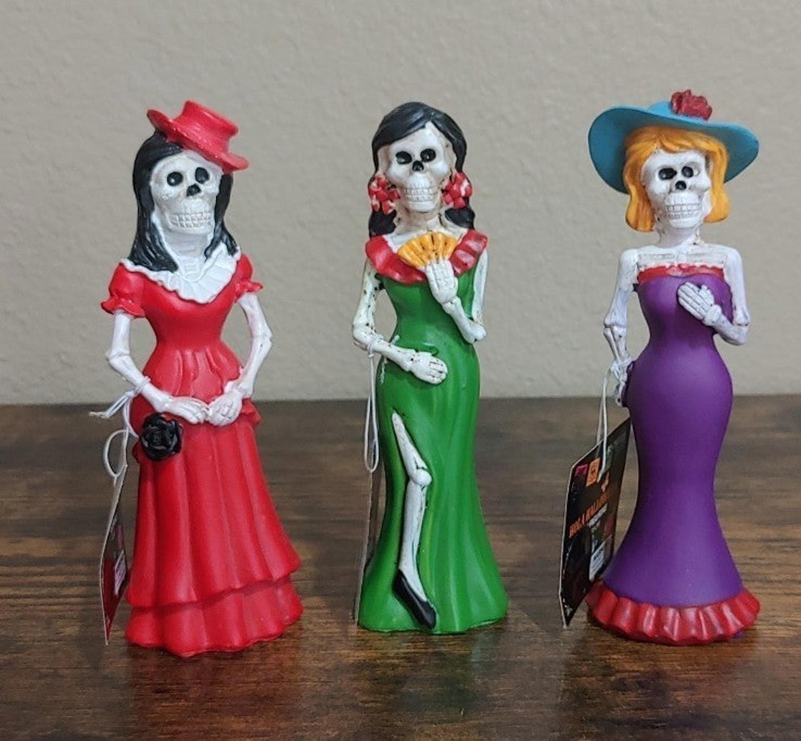Three Catrina figurines with red, green, and purple dresses