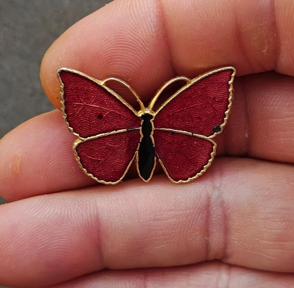 Red butterfly enamel brooch with gold-tone outline and pin, close-up on fingers.